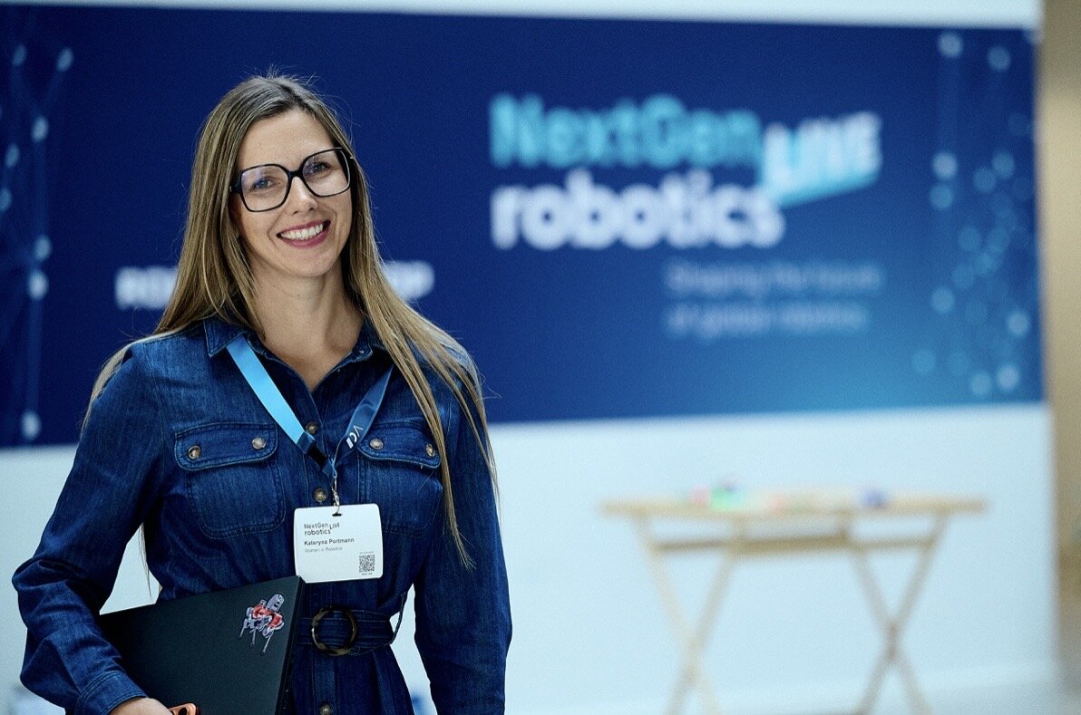 Kateryna Portmann walking at the NextGen Robotics Live event, holding a folder and wearing a name badge